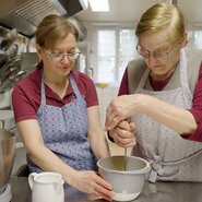 Zwei Frauen in Schürzen kochen oder backen zusammen in einer Küche, eine prüft die Zutaten in einer Schüssel, die andere rührt in einem Topf. | © Urlaub am Bauernhof / Daniel Gollner