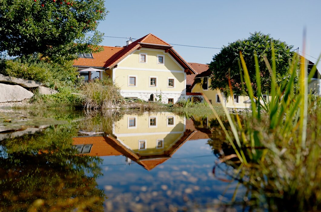 Ein idyllisches Landhaus mit Holzdach spiegelt sich in einem friedlichen Teich, umgeben von üppiger Vegetation und klarem Himmel. Die Atmosphäre vermittelt Ruhe und Naturnähe.