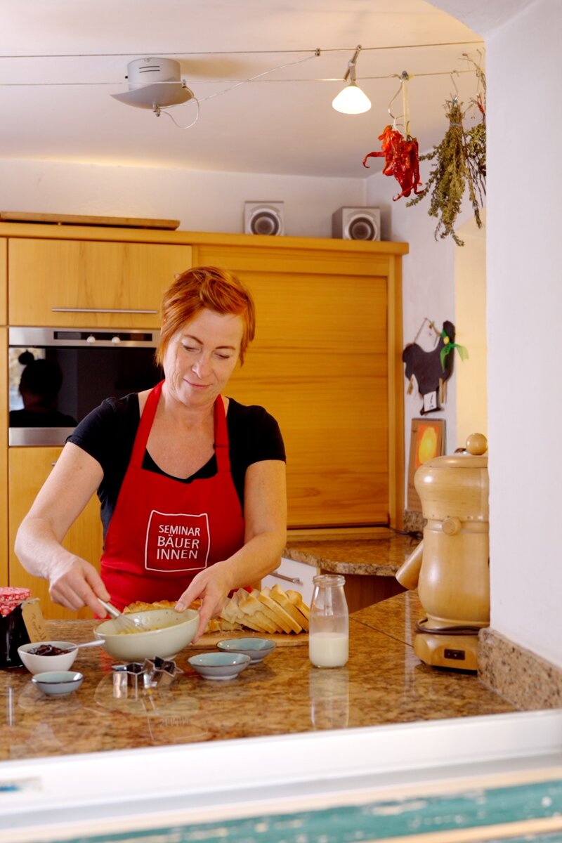 Die Person in einer roten Schürze bereitet Backwaren zu, umgeben von einer gemütlichen, dekorativen Küche mit hellen Holzelementen. | © Urlaub am Bauernhof / Daniel Gollner