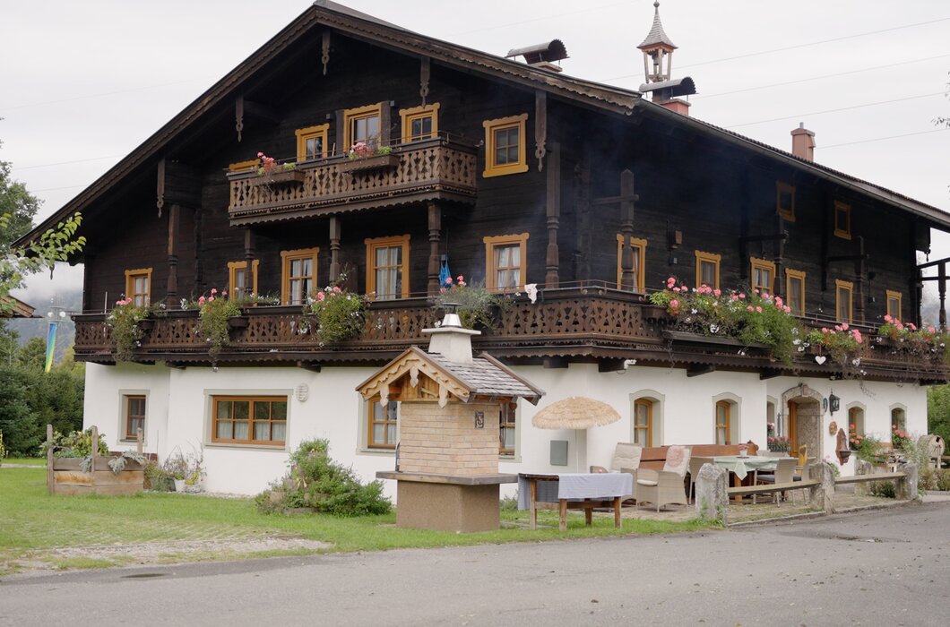 Traditionelles bayerisches Landhaus mit Holzfassade, großem Balkon und Blumenkästen, umgeben von Grünfläche und Bäumen. Sitzgelegenheiten und Gartendekoration deuten auf einen touristischen Betrieb hin. | © Urlaub am Bauernhof / Daniel Gollner