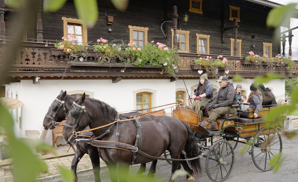 Eine historische Kutsche mit Pferdegespann fährt an einem mit Blumen geschmückten, traditionellen Holzhaus vorbei, in dem Gäste auf den Balkonen sitzen. | © Urlaub am Bauernhof / Daniel Gollner