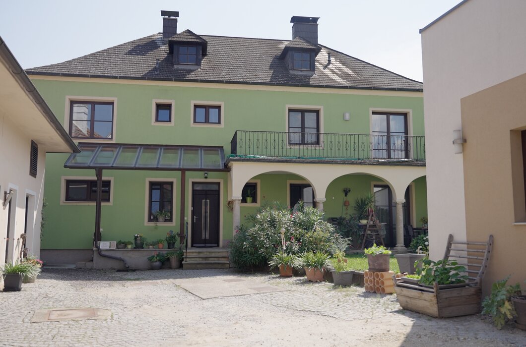 Zweistöckiges Haus mit grüner Fassade, Balkonen und Arkaden. Gepflasterter Hof mit Pflanzen und Gartendekoration. Historisch anmutendes Gebäude in ruhiger Atmosphäre. | © Urlaub am Bauernhof / Daniel Gollner