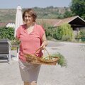 Eine Frau mittleren Alters in einem roten Punktekleid hält einen Korb mit Kräutern auf einem ländlichen Grundstück, umgeben von Garten und Wohnhäusern. | © Urlaub am Bauernhof / Daniel Gollner