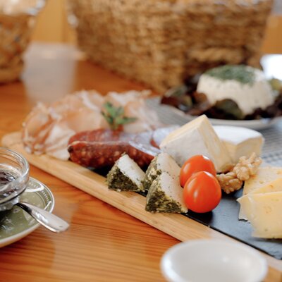 Verschiedene Käsesorten, Wurst, Pesto und Gebäck auf einem Holzbrett, eine Tasse Essig oder Sirup, Tomaten und weitere Delikatessen - ein vielfältiges, appetitliches Frühstücks- oder Snack-Arrangement. | © Urlaub am Bauernhof / Daniel Gollner