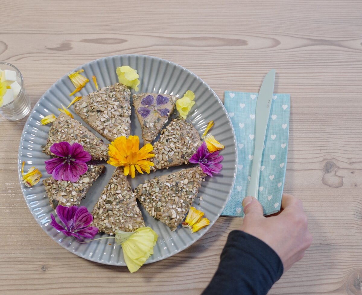 Teller mit verschiedenen Vollkornbrötchen, dekoriert mit bunten Blumen und Blättern, auf Holztisch mit blauer, gepunkteter Serviette. | © Urlaub am Bauernhof / Daniel Gollner