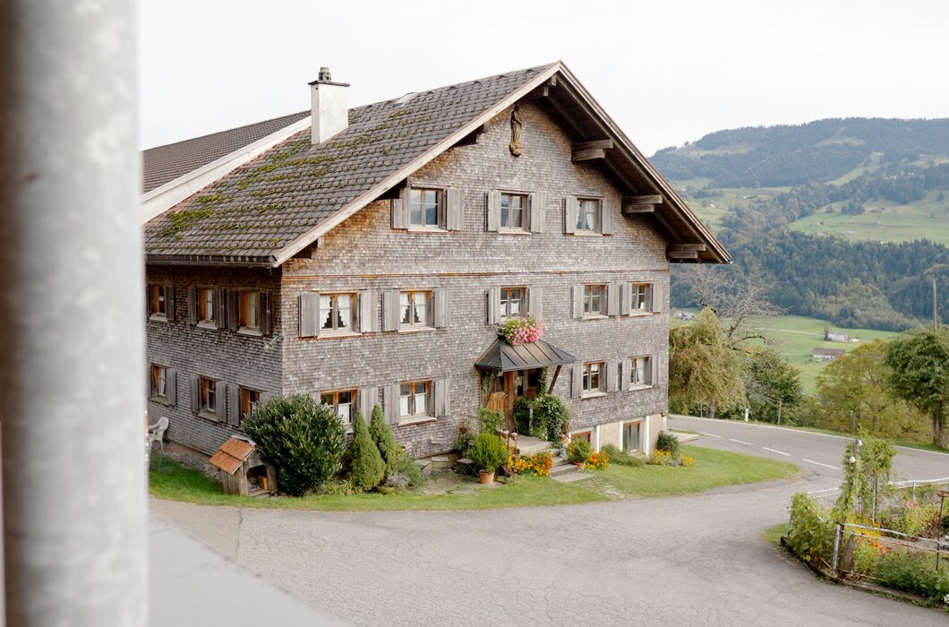 Traditionelles zweistöckiges Holzhaus mit Satteldach, umgeben von Natur und dekoriert mit Blumen. Es befindet sich auf einer Erhebung an einer Straße, mit Wiesen und Bergen im Hintergrund. | © Urlaub am Bauernhof / Daniel Gollner