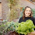 Eine freundlich lächelnde Frau hält einen großen Salatkopf, umgeben von Blumen und einer ziegelsteinernen Wand. | © Urlaub am Bauernhof / Daniel Gollner
