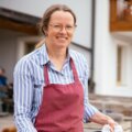Eine Frau in Schürze präsentiert stolz ein frisch gebackenes Stück Brot vor einem Berghotel-Gebäude mit Balkonen und Holzelementen.