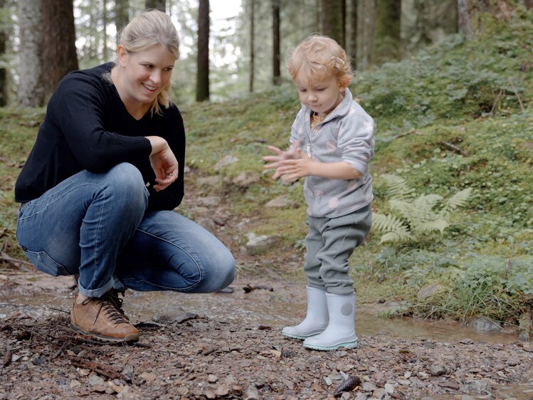 Eine lächelnde Frau beobachtet ein kleines Kind, das auf dem Waldboden spielt. Beide tragen passende Outdoorkleidung und Gummistiefel. Die natürliche Umgebung und Vegetation im Hintergrund lassen auf einen Spaziergang in einem Waldgebiet schließen. | © Urlaub am Bauernhof / Daniel Gollner