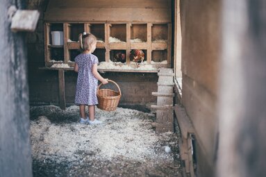 Ein kleines Mädchen in einem Kleid steht in einem alten Bauernhofstall voller Holzregale und Körbe. Sie hält einen Korb in der Hand und blickt in Richtung der Kamera. | © Urlaub am Bauernhof / Daniel Gollner
