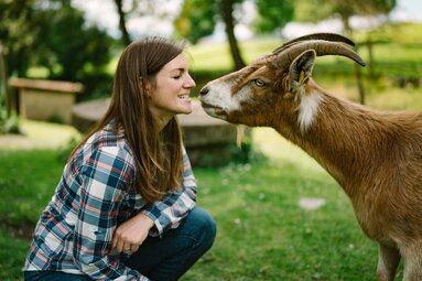 Eine lächelnde Frau in einem karierten Hemd interagiert liebevoll mit einem braunen Ziegenbock in einer idyllischen Umgebung voller Bäume und Wiesen. | © Urlaub am Bauernhof / Daniel Gollner 