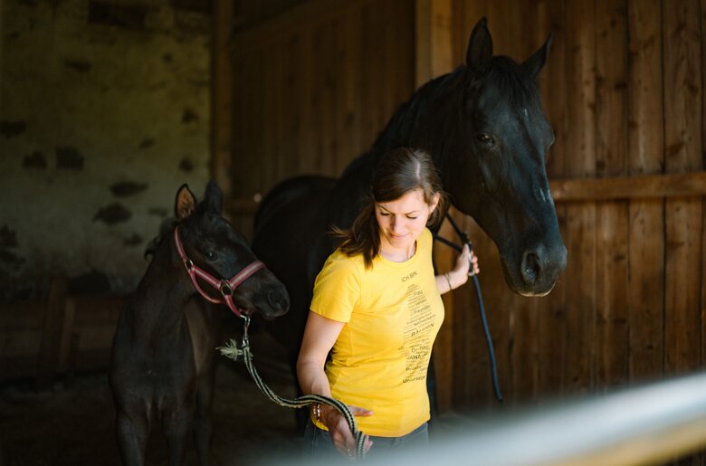 Eine junge Frau in einem gelben Shirt steht zwischen zwei schwarzen Pferden in einem hölzernen Stall. Sie lächelt und hält eines der Pferde an der Leine. | © Urlaub am Bauernhof / Daniel Gollner 