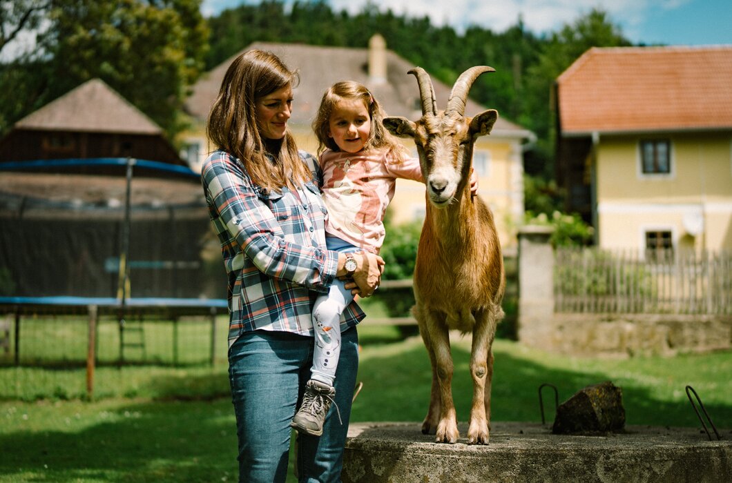 Eine Frau hält ein kleines Mädchen auf dem Arm, während neben ihnen eine Ziege auf einer Erhöhung steht. Im Hintergrund sind Häuser und Bäume zu sehen, die eine ländliche Umgebung andeuten. | © Urlaub am Bauernhof / Daniel Gollner