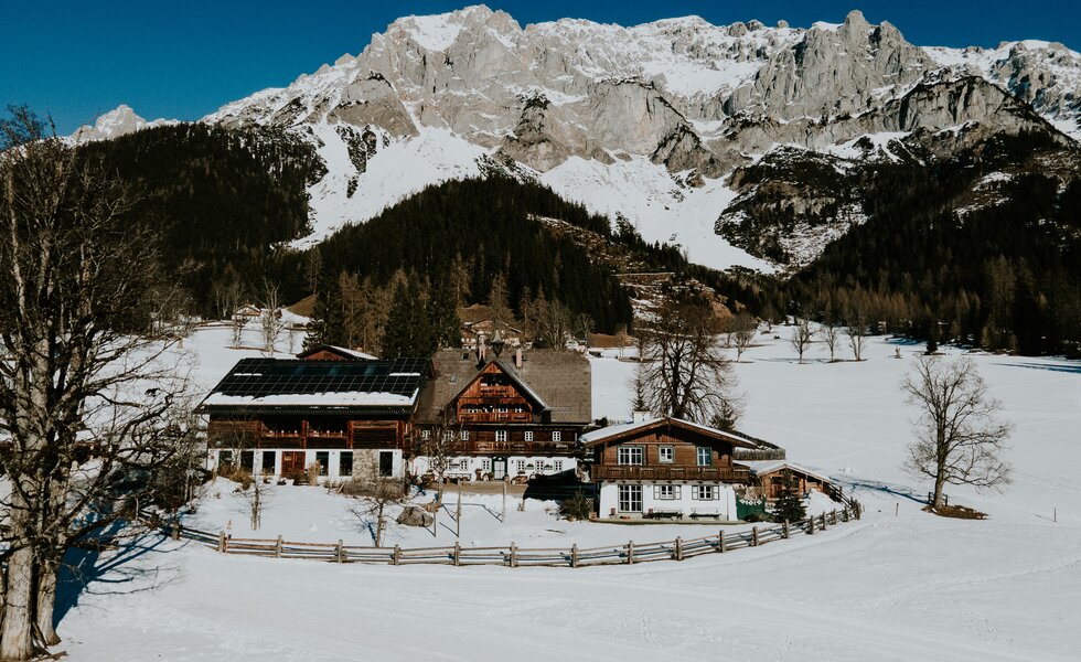Traditionelles Bergdorf inmitten verschneiter Berglandschaft, umgeben von Wälder und einem Zaun. Mehrere Holzhäuser mit typischer Architektur sind zu sehen, einige mit Solarpanelen auf den Dächern. | © Urlaub am Bauernhof / Daniel Gollner