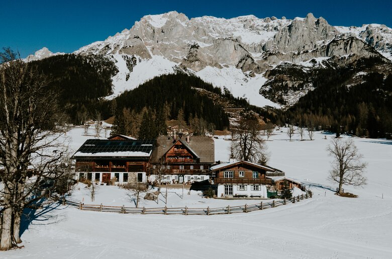 Traditionelles Bergdorf inmitten verschneiter Berglandschaft, umgeben von Wälder und einem Zaun. Mehrere Holzhäuser mit typischer Architektur sind zu sehen, einige mit Solarpanelen auf den Dächern. | © Urlaub am Bauernhof / Daniel Gollner