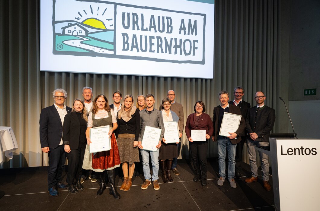 Eine Gruppe von Personen, einige in traditioneller Tracht, stehen auf einer Bühne und halten Urkunden. Im Hintergrund ist ein grafisches Logo eines landwirtschaftlichen Betriebs zu sehen. | © Urlaub am Bauernhof / Christian Lendl