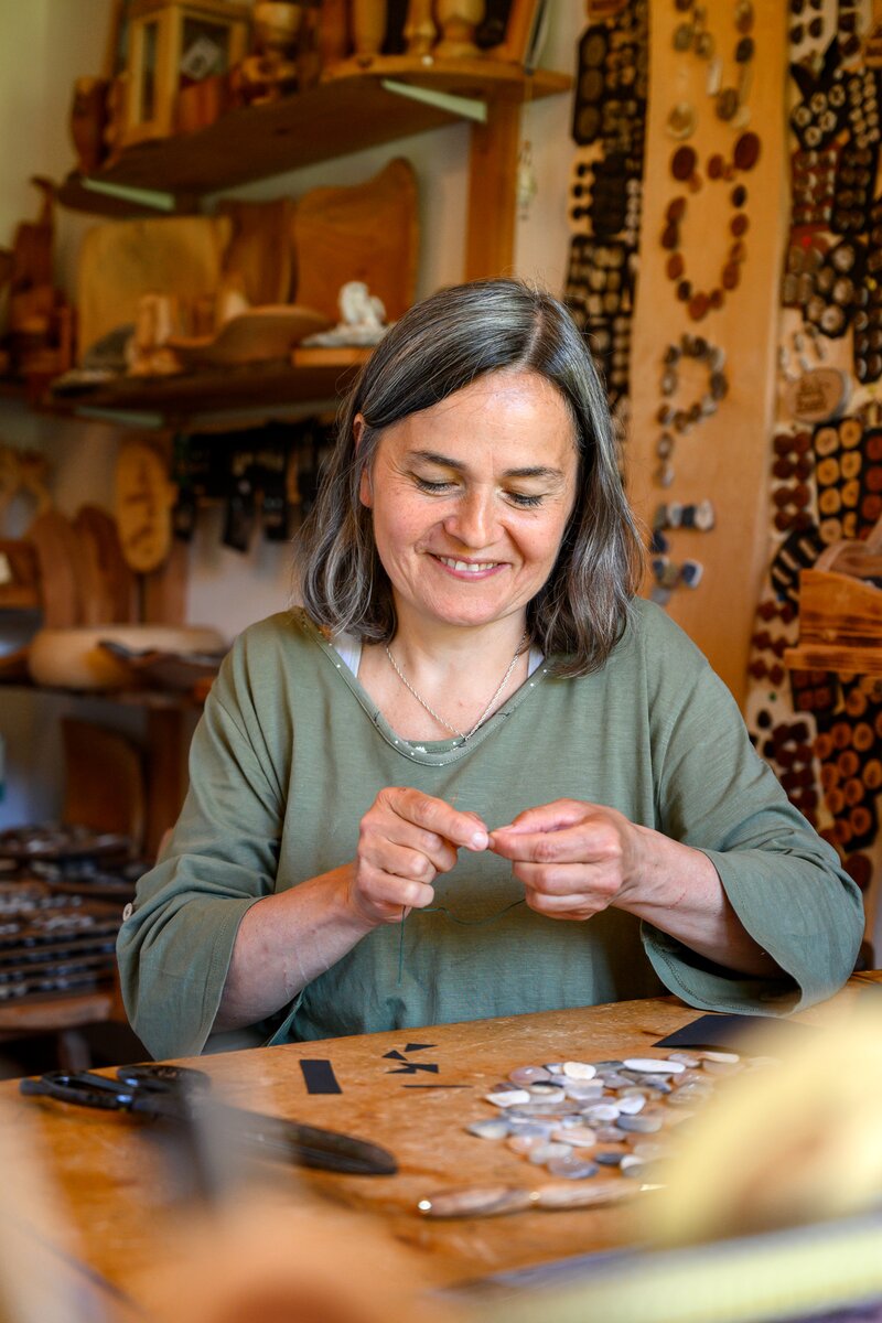 Eine Frau sitzt lächelnd an einem Holztisch voller Schmuckstücke und hantiert damit, umgeben von Regalen mit dekorativen Gegenständen in einem gemütlichen Ambiente. | © Urlaub am Bauernhof / Punkt und Komma