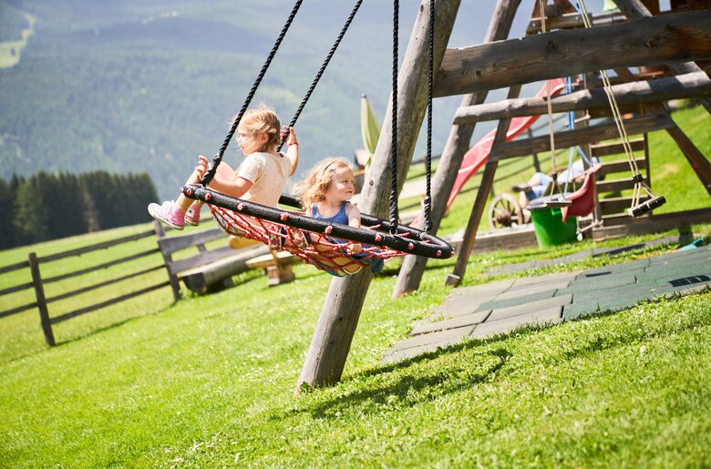 Zwei Mädchen schaukeln auf einer bunten, hölzernen Schaukel vor einem ländlichen Hintergrund mit Wiese, Zaun und Bergen. | © Urlaub am Bauernhof / Punkt und Komma