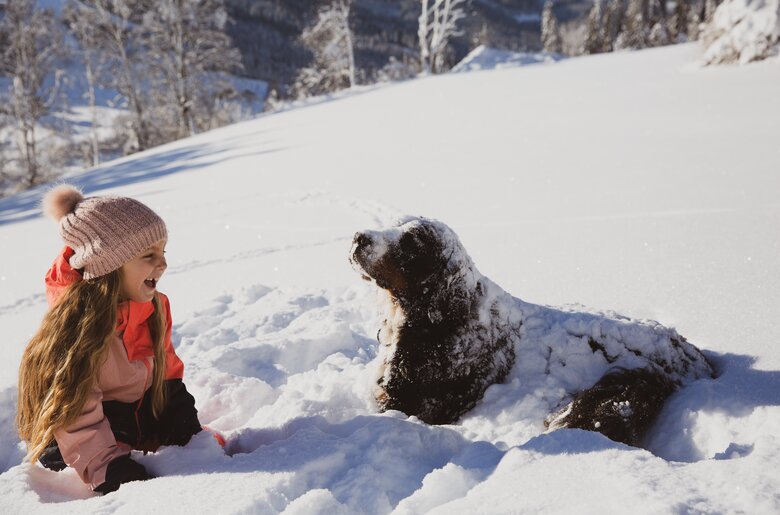 Ein Mädchen in roter Jacke sitzt im Schnee und spielt mit einem schwarzen Hund. Umgeben von schneebedeckten Bäumen in einer winterlichen Landschaft. | © Urlaub am Bauernhof / Pascal Baronit