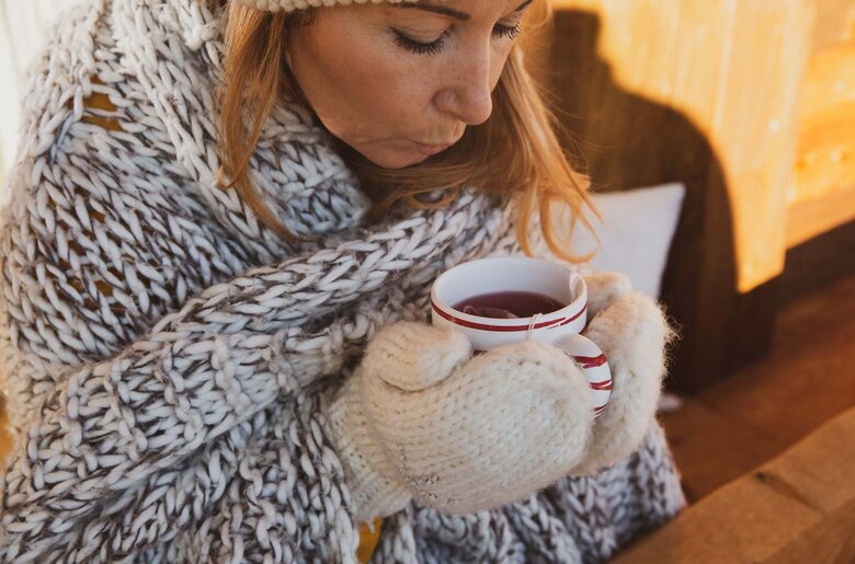 In einem warmen Ambiente sitzt eine Person in einem gemütlichen, gestrickten Schal und genießt entspannt eine Tasse Tee. Die Stimmung ist gemütlich und einladend. | © Urlaub am Bauernhof / Pascal Baronit