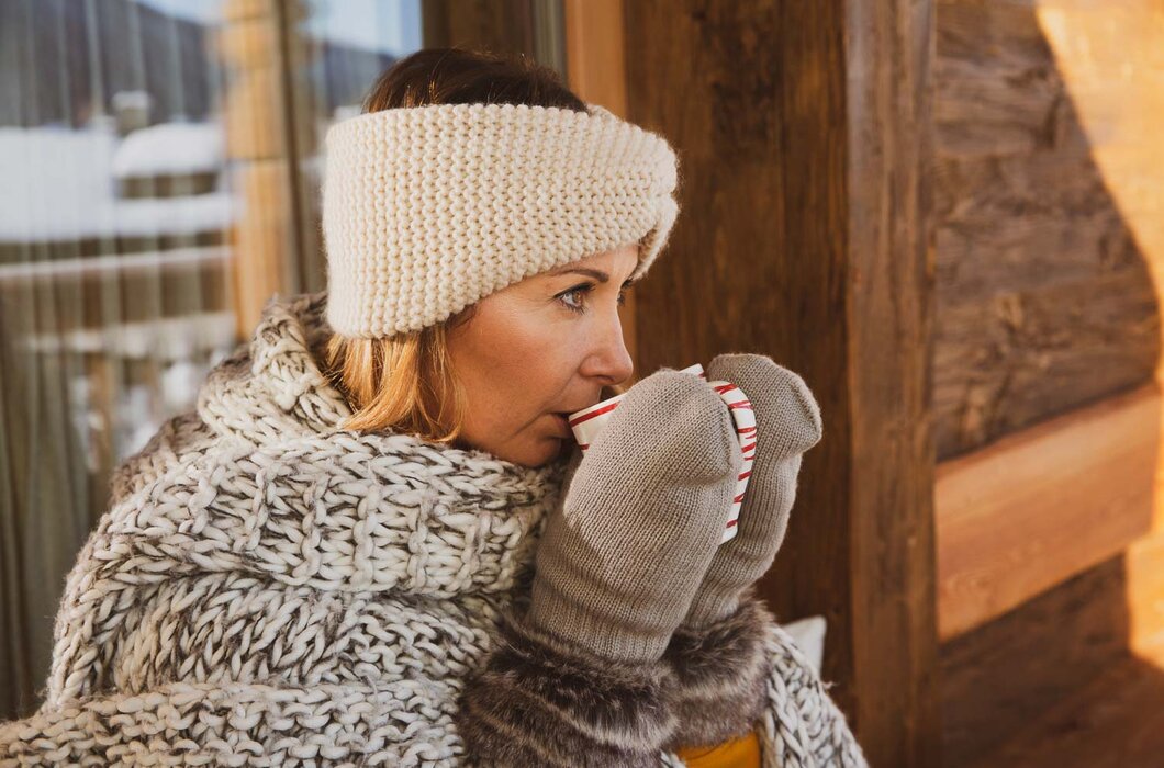 Eine Frau in warmer, gemusterter Kleidung und Mütze trinkt einen heißen Getränk, während sie sich an eine Holzwand lehnt. | © Urlaub am Bauernhof / Pascal Baronit