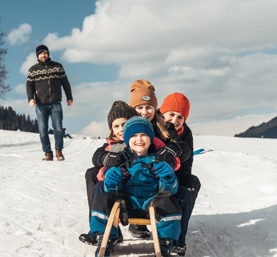Eine Familie genießt einen winterlichen Ausflug: Ein Kind sitzt in einem Schlitten, von Erwachsenen umgeben, die mit warmer Kleidung bekleidet sind. Im Hintergrund eine schneebedeckte Landschaft und Wolken am Himmel. | © Urlaub am Bauernhof / Punkt und Komma