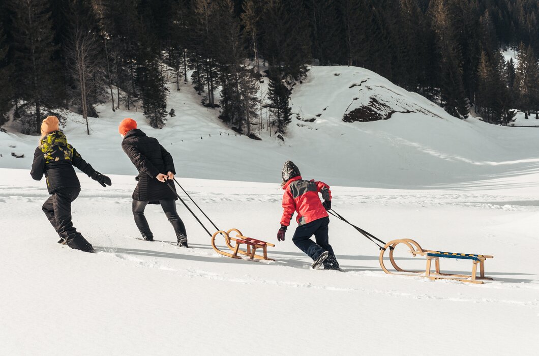Drei Personen ziehen Schlitten über verschneite Landschaft mit bewaldeten Hügeln im Hintergrund. | © Urlaub am Bauernhof / Punkt und Komma