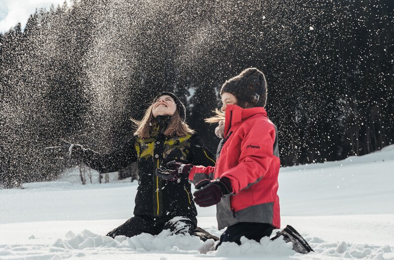 Zwei fröhliche Personen spielen im verschneiten Winterwald, in dem die Schneeflocken in der Luft tanzen. Sie scheinen die Natur und die Winterlandschaft zu genießen. | © Urlaub am Bauernhof / Punkt und Komma