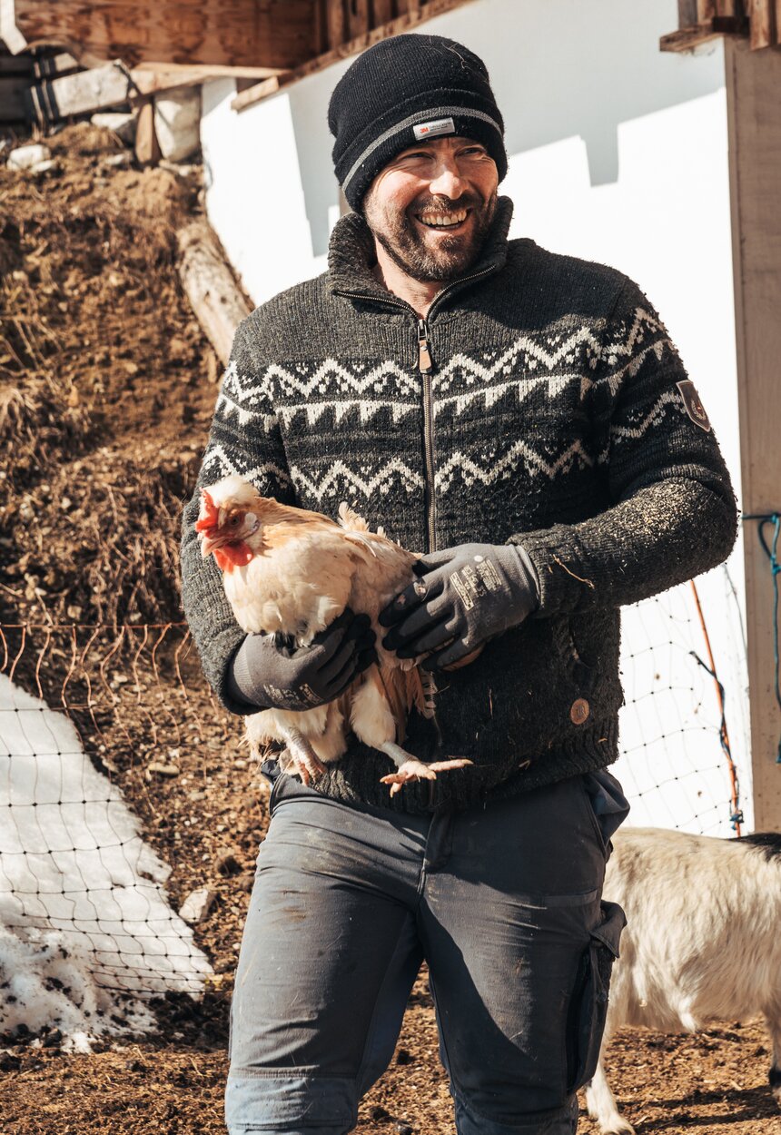 Mann in Winterpullover hält Henne auf der Veranda einer Holzhütte. Umgeben von Schnee und Holz. | © Urlaub am Bauernhof / Punkt und Komma