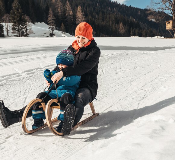 Eine Frau in orangefarbener Mütze und dunkler Jacke rodelt auf einer verschneiten Piste, eng umschlungen mit einem Kind in blauer Winterkleidung. Das Lächeln der Frau und die Schneelandschaft verleihen der Szene eine fröhliche, winterliche Stimmung. | © Urlaub am Bauernhof / Punkt und Komma