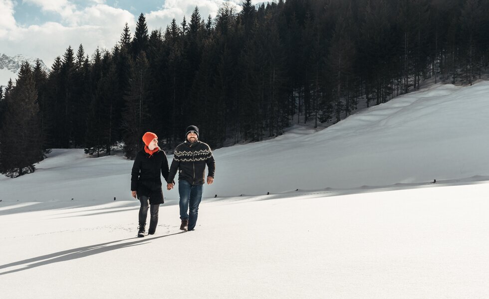 Zwei Personen gehen Händchen haltend durch eine winterliche Landschaft mit Schneehügeln und verschneiter Berglandschaft im Hintergrund. | © Urlaub am Bauernhof / Punkt und Komma