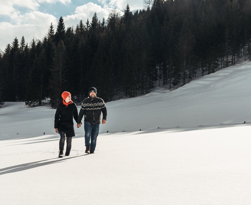 Zwei Personen gehen Händchen haltend durch eine winterliche Landschaft mit Schneehügeln und verschneiter Berglandschaft im Hintergrund. | © Urlaub am Bauernhof / Punkt und Komma