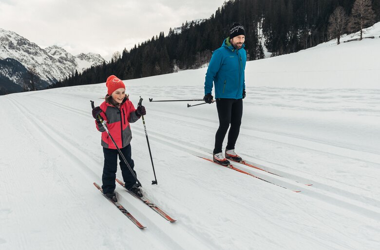 Eine Person in Winterbekleidung, die Ski fährt, und eine weitere Person neben ihr, ebenfalls Ski fahrend, in einer schneebedeckten Winterlandschaft mit Bergen im Hintergrund. | © Urlaub am Bauernhof / Punkt und Komma