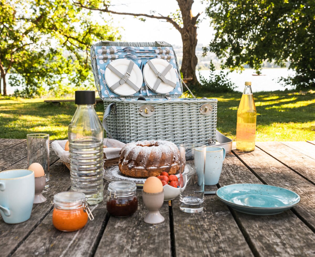 Picnic basket, cake, strawberries, drinks, and tableware on wooden table in garden with lush greenery. | © Urlaub am Bauernhof / Punkt und Komma