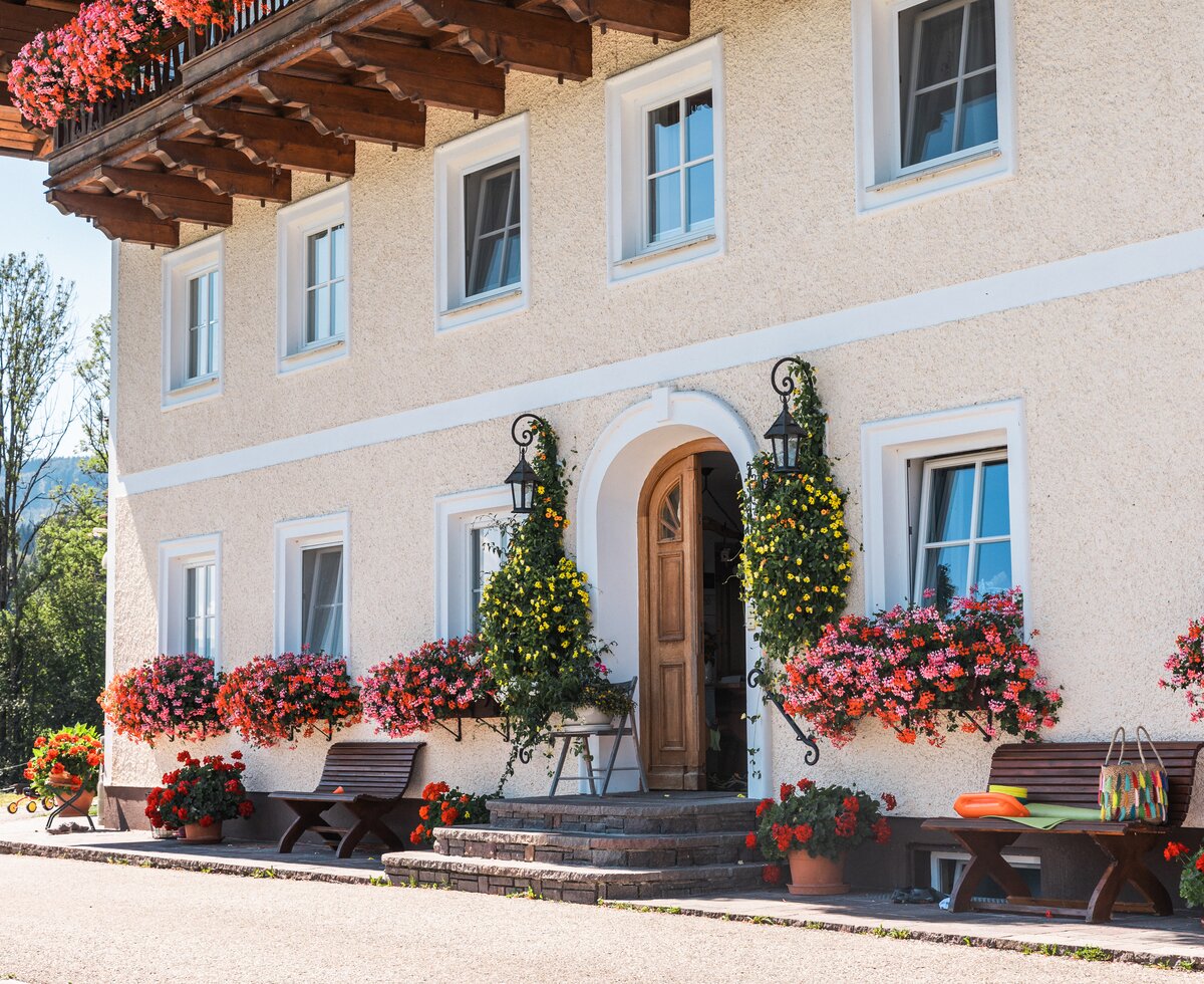 Picturesque rustic building with wooden beams, colorful flower boxes, and ornate entrance decorated with potted plants. | © Urlaub am Bauernhof / Punkt und Komma