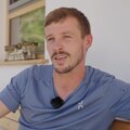 Der lächelnde Mann mit Bart und mittellanger Frisur trägt ein blaues Shirt mit markanter Applikation.
