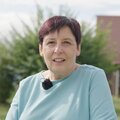 Eine Frau mittleren Alters mit kurzen dunklen Haaren steht in einer ländlichen Umgebung und lächelt in die Kamera. Sie trägt einen hellblauen Pullover und einen dunklen Schal.