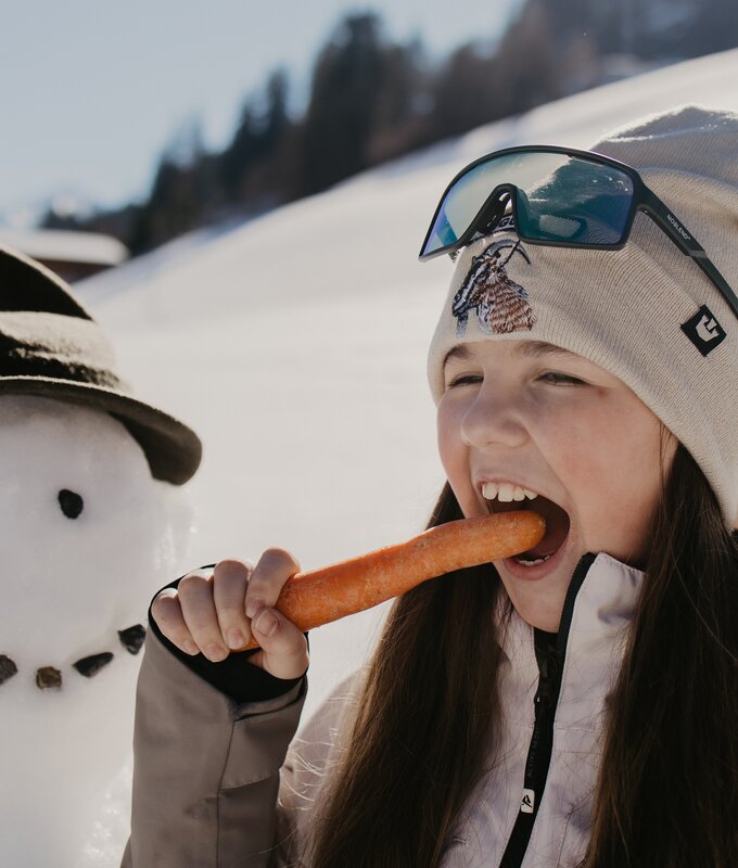 Ein Mädchen beißt in die Karotte vom Schneemann der neben ihr steht. | © Urlaub am Bauernhof im SalzburgerLand/ Love_Lens_Photograph