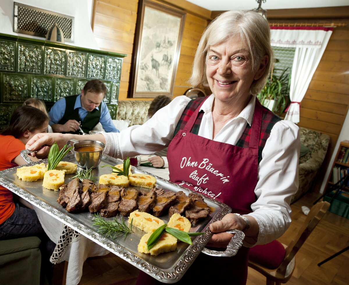 Bäuerin zeigt Fleischplatte, Hochsteiermark, Steiermark | © Urlaub am Bauernhof Steiermark / Werner Krug Die Bäuerin zeigt die Fleischplatte mit Familie im Hintergrund, Hochsteiermark, Steiermark | © Urlaub am Bauernhof Steiermark / Werner Krug