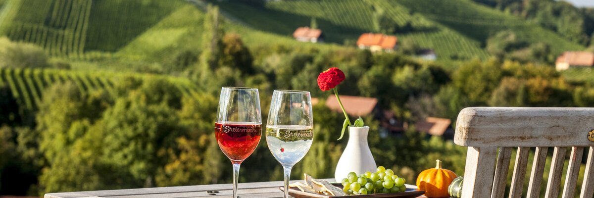 Weingläser stehen auf einem Holztisch, typisch südsteirische Weinhänge im Hintergrund, Süd-West-Steiermark, Steiermark | © Süd-West-Steiermark / ikarus.cc