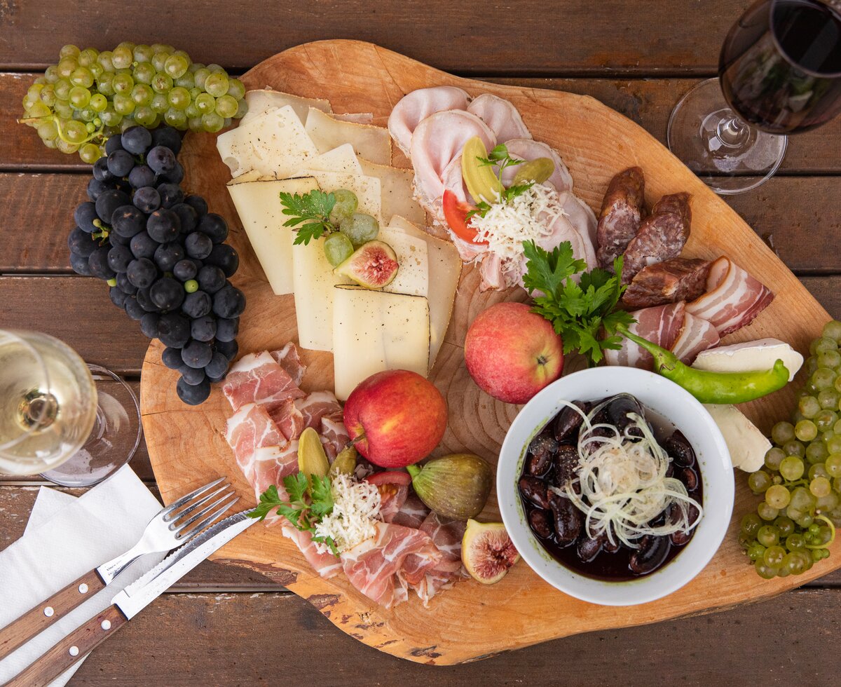 Eine herzhafte steirische Brettljause am Holzbrett serviert, mit Würsteln, Speck, Rohschinken, Käse, Weintrauben, Äpfeln, frischen Feigen und Käferbohnensalat | © Urlaub am Bauernhof / punkt & komma