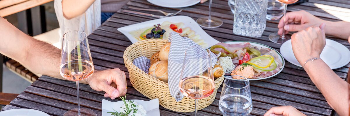Eine Gruppe genießt ein luxuriöses Picknick mit Köstlichkeiten auf einer Terrasse. Auf dem Holztisch stehen Weinkelche, eine Brotkorb und eine kreative Präsentation von Appetithäppchen.