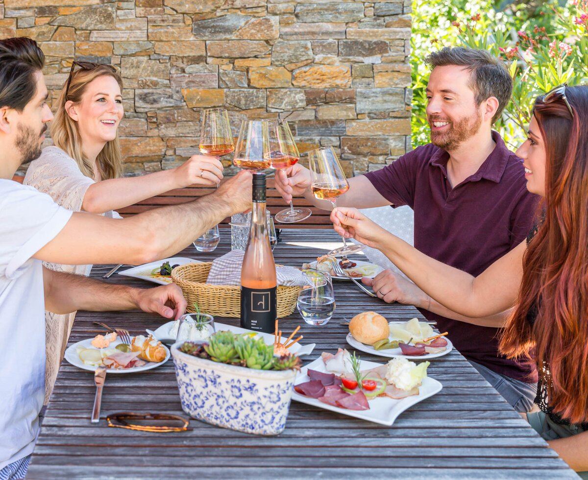 Eine Gruppe von Freunden genießt zusammen ein Picknick im Freien. Sie sitzen an einem Holztisch, essen verschiedene Snacks und Getränke und stoßen mit Weingläsern an. Die Szenerie wird von einer Natursteinwand und üppiger Vegetation umrahmt, was eine gemütliche Atmosphäre schafft.