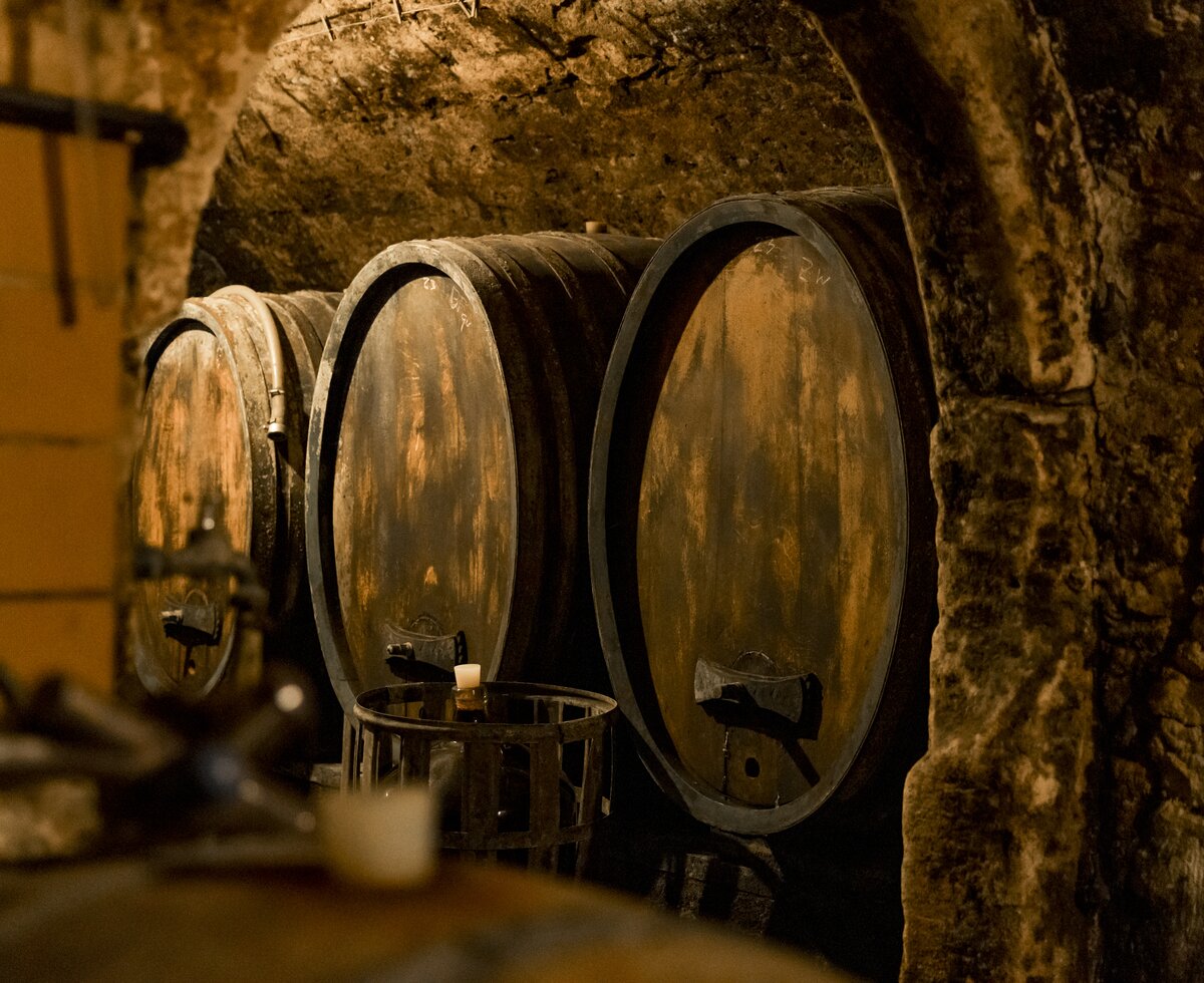 Alte, abgenutzte Holzfässer in einem düsteren Keller, wobei ihr Inhalt nicht erkennbar ist. Die Fässer sind in einer Reihe aufgestellt und teilweise von Steinmauern umgeben, wodurch eine rustikale, authentische Atmosphäre entsteht.