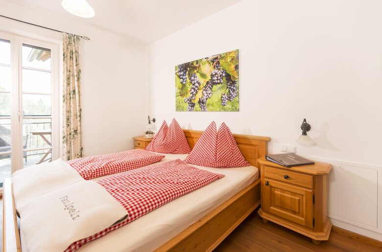 Gemütliches Hotelzimmer mit Holzmöbeln, Karo-Bettwäsche in Rot und Weiß sowie einem Bild von Trauben an der Wand. Der Blick geht durch ein Fenster auf eine Terrasse.