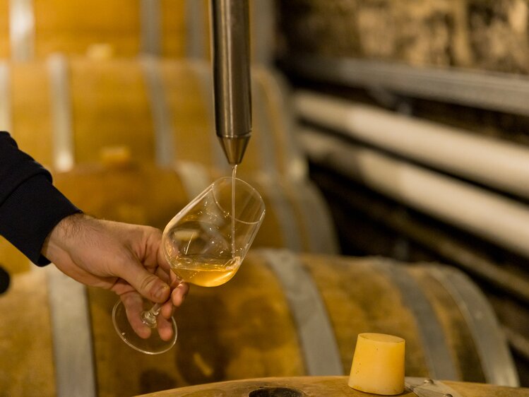 Eine Person in einem schwarzen Anzug gießt Whisky in ein Glas, umgeben von Fässern in einem Weinkeller.