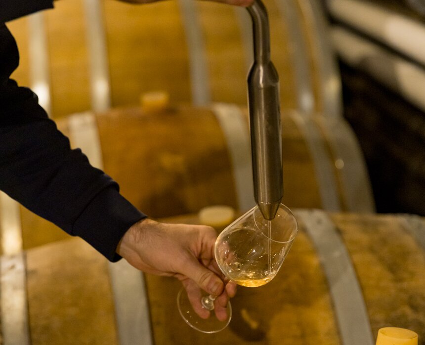 Eine Person in dunklem Anzug probiert den Inhalt aus einem Glas, das sich in einem Holzfass befindet. Im Hintergrund sind weitere Fässer zu sehen.