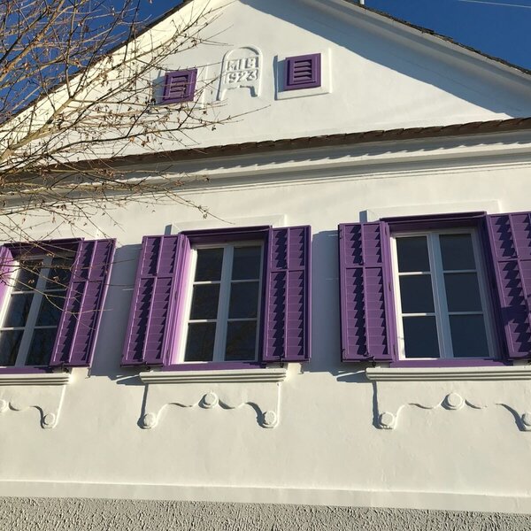 The white facade of the Farm House features three windows with purple shutters and decorative elements, including the year 1923 on the gable.