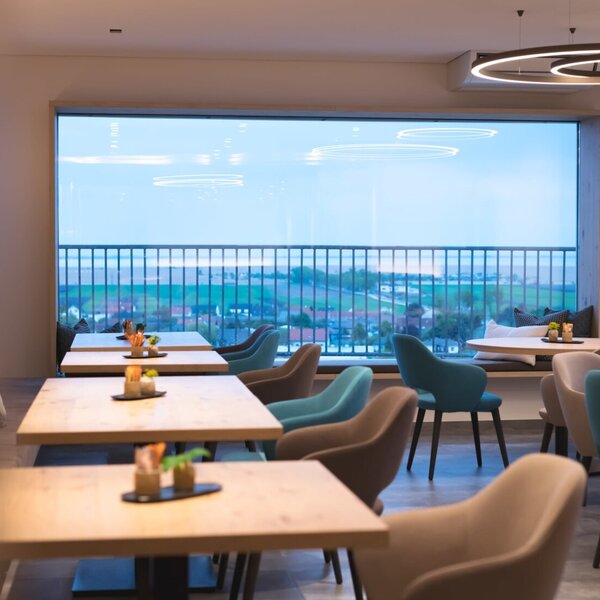 Der Aufenthaltsraum des Hofes mit Tischen und Stühlen und einer großen Fensterfront, die einen weiten Blick auf die Umgebung bietet.