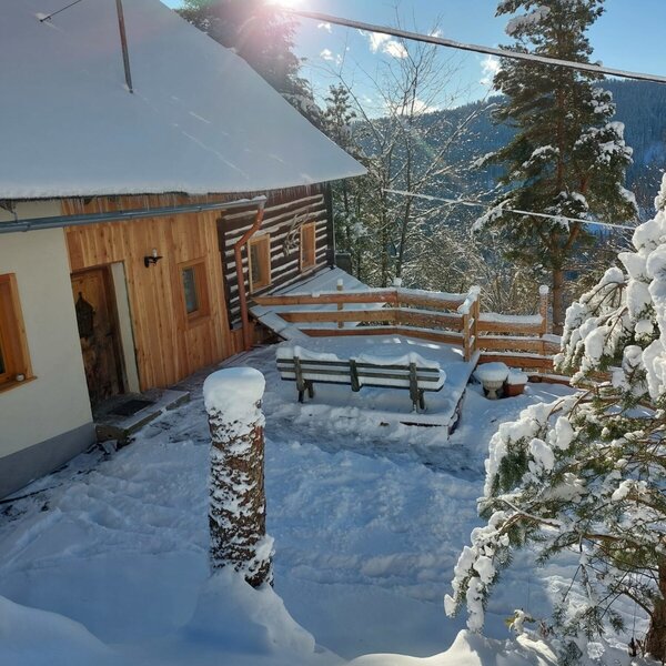 The Alpine Hut's snow-covered exterior and wooden terrace with a bench, set against a backdrop of snowy mountains and forests.